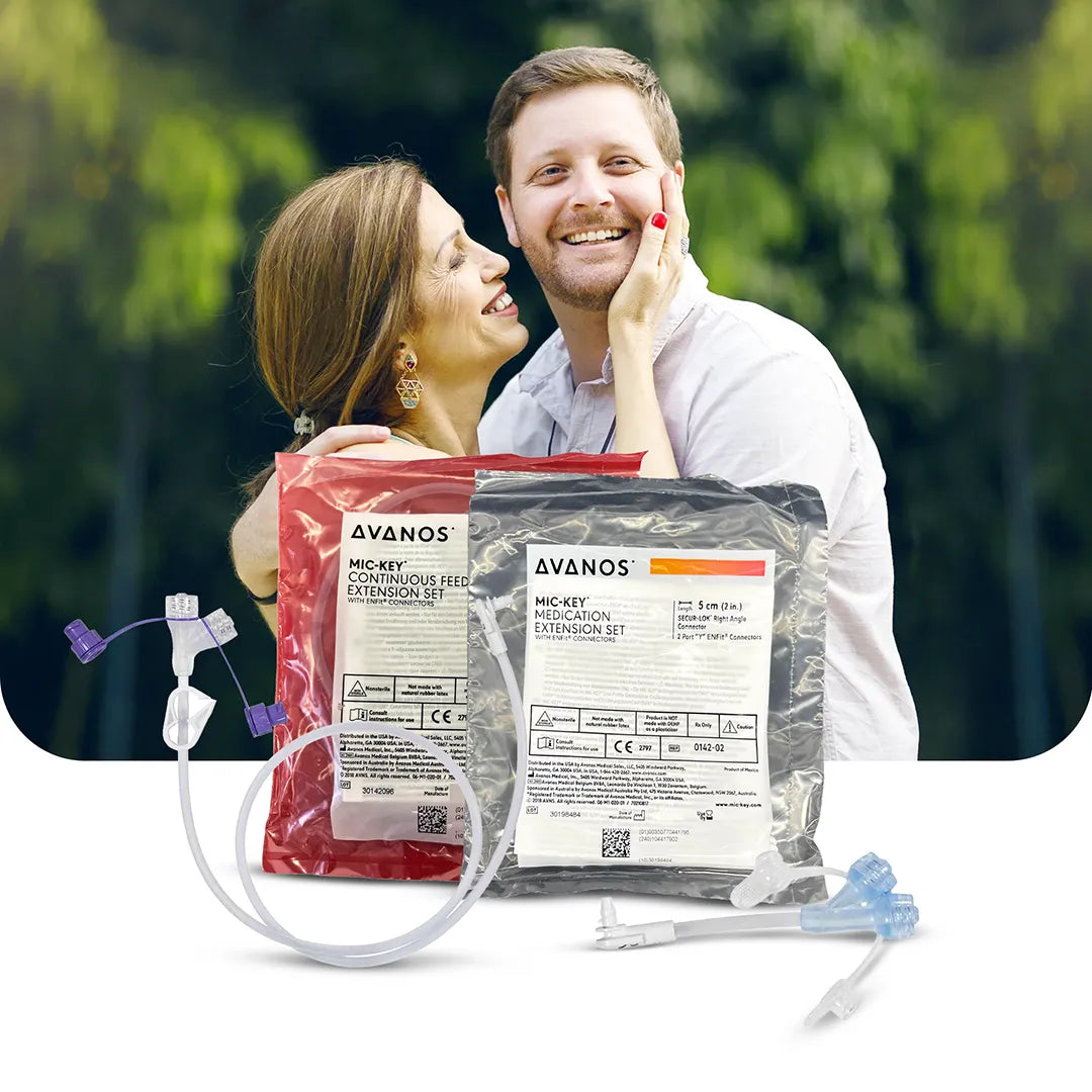 Smiling couple with Avanos MIC-KEY Medication and Continuous Feed Extension Sets in foreground, featuring ENFit-compatible connectors, available at Medisa.